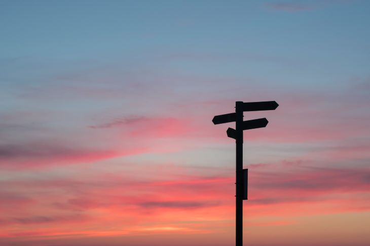 A silhouetted signpost with multiple arrows against a pink sunset sky