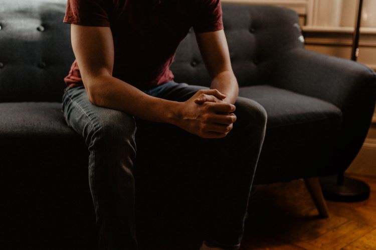 The lower half of a man with his hands clasped, sat on a sofa